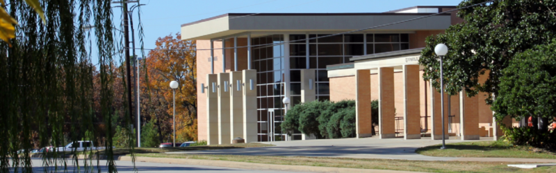 Modern brick and glass building with tall windows by a tree-lined road, blue sky above and autumn trees in the background.