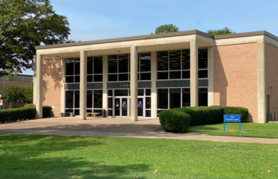 Two-story brick library with large front windows, green lawn, trimmed bushes, blue sign near entrance, and trees around.