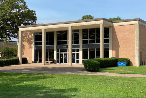 Two-story brick library with large front windows, green lawn, trimmed bushes, blue sign near entrance, and trees around.