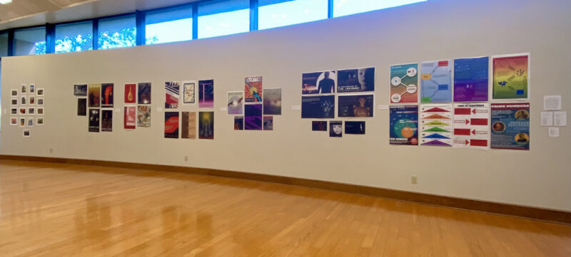 Gallery with wooden floors, colorful posters and art on a white wall, large windows above, and bright ceiling lights.