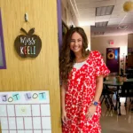 AAT AC Alumni at SFASU Charter Bright Smiling woman in red and white floral dress stands by classroom door labeled Miss Keeli; tables, chairs, and Jot Lot calendar inside.