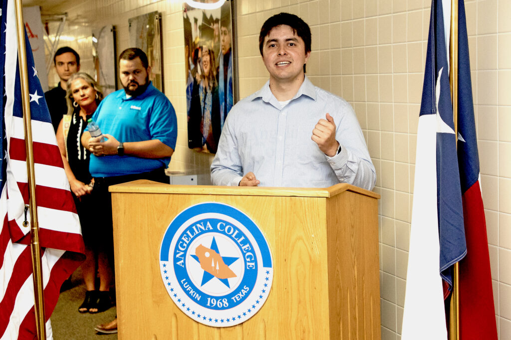 Man speaks at Angelina College podium between U.S. and Texas flags; three people listen in the hallway behind him.