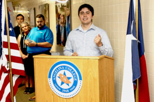 Man speaks at Angelina College podium between U.S. and Texas flags; three people listen in the hallway behind him.