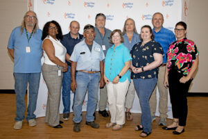 Ten adults smile for a group photo indoors before an Angelina College backdrop, casually dressed on a wooden floor.
