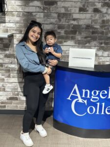Woman smiling and holding a child beside a blue counter with part of the Angels College logo and a boxed iPad; gray brick wall behind.