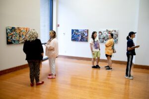 Five people observe colorful abstract paintings in a bright art gallery; some talk in pairs, others look at artwork alone.