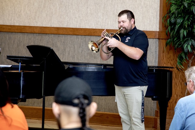 Man in a black shirt plays a brass instrument by a grand piano, performing for an audience; large plant in the background.