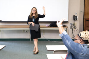 Woman in black dress smiles and waves at front of classroom, holding folder. Student in foreground raises hand. Blank screen behind.