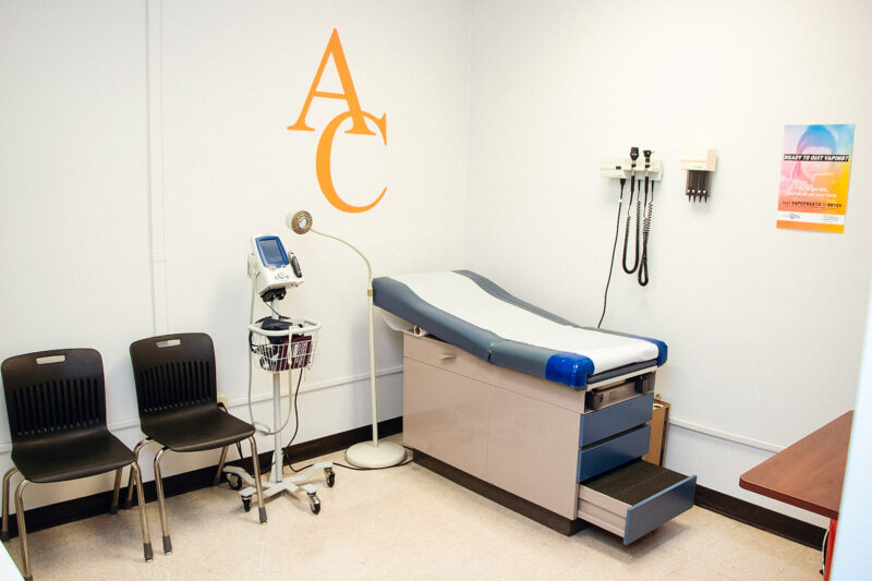 Small, tidy exam room with table, two black chairs, wall-mounted medical equipment, rolling monitor, and "AC" in orange above table.