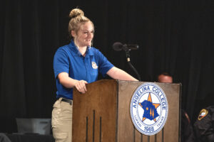 Woman in a blue shirt speaks into a microphone at an Angelina College podium on a stage with a black background.