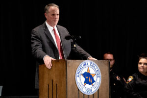A suited man speaks at a podium with the Angelina College seal; uniformed people sit behind him on a dimly lit stage.