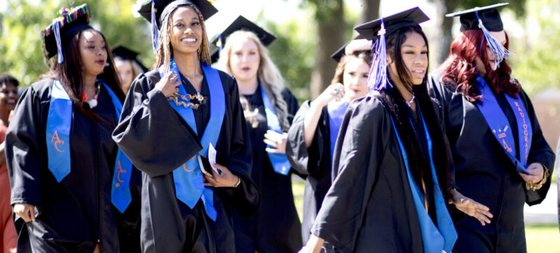Graduates in black caps, gowns, and blue stoles walk outside, smiling amid trees and sunlight.