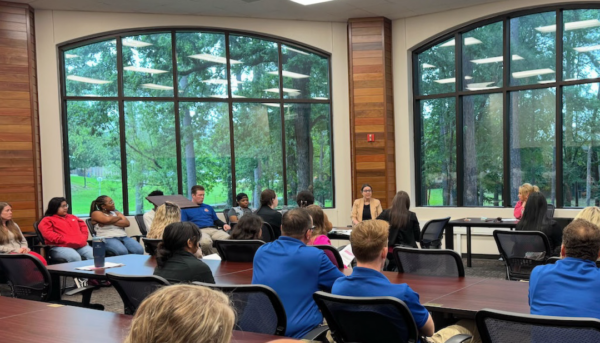 People seated in a conference room with large windows listen to two speakers; wooden accents and trees visible outside.