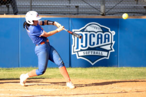 Softball player in blue uniform and white helmet swings bat, hitting ball; NJCAA Softball logo visible on fence behind.