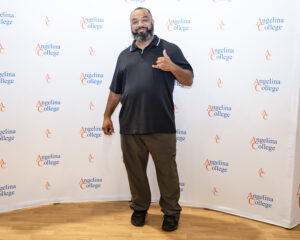 Smiling man in black polo and gray pants stands on wood floor before Angelina College logo backdrop, making a shaka gesture.