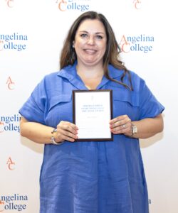 Woman in a blue dress smiles, holding a plaque in front of an Angelina College backdrop.