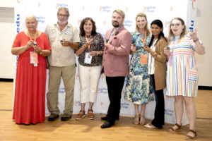 Seven adults, men and women in business and casual attire, smile holding small items before an Angelina College backdrop indoors.