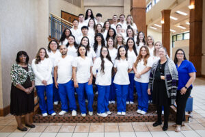 Nursing students in white tops and blue pants pose on stairs with four professionally dressed smiling instructors indoors.