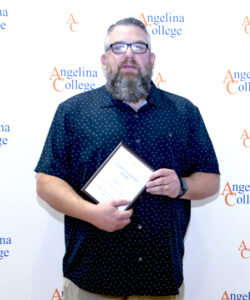 Man with glasses in a dark patterned shirt holds a plaque in front of an Angelina College logo step-and-repeat banner.
