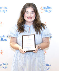 Smiling woman with long brown hair in a light blue striped dress holds a plaque before an Angelina College banner.