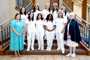 Nurses in white uniforms and caps pose on stairs with two women in different attire: a turquoise dress and a navy cape indoors.