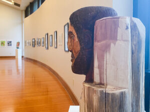 Wooden sculpture of a man's head on a pedestal in an art gallery with polished floors and framed art along a curved wall.
