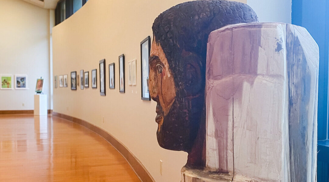 Wooden sculpture of a man's head on a pedestal in an art gallery with polished floors and framed art along a curved wall.