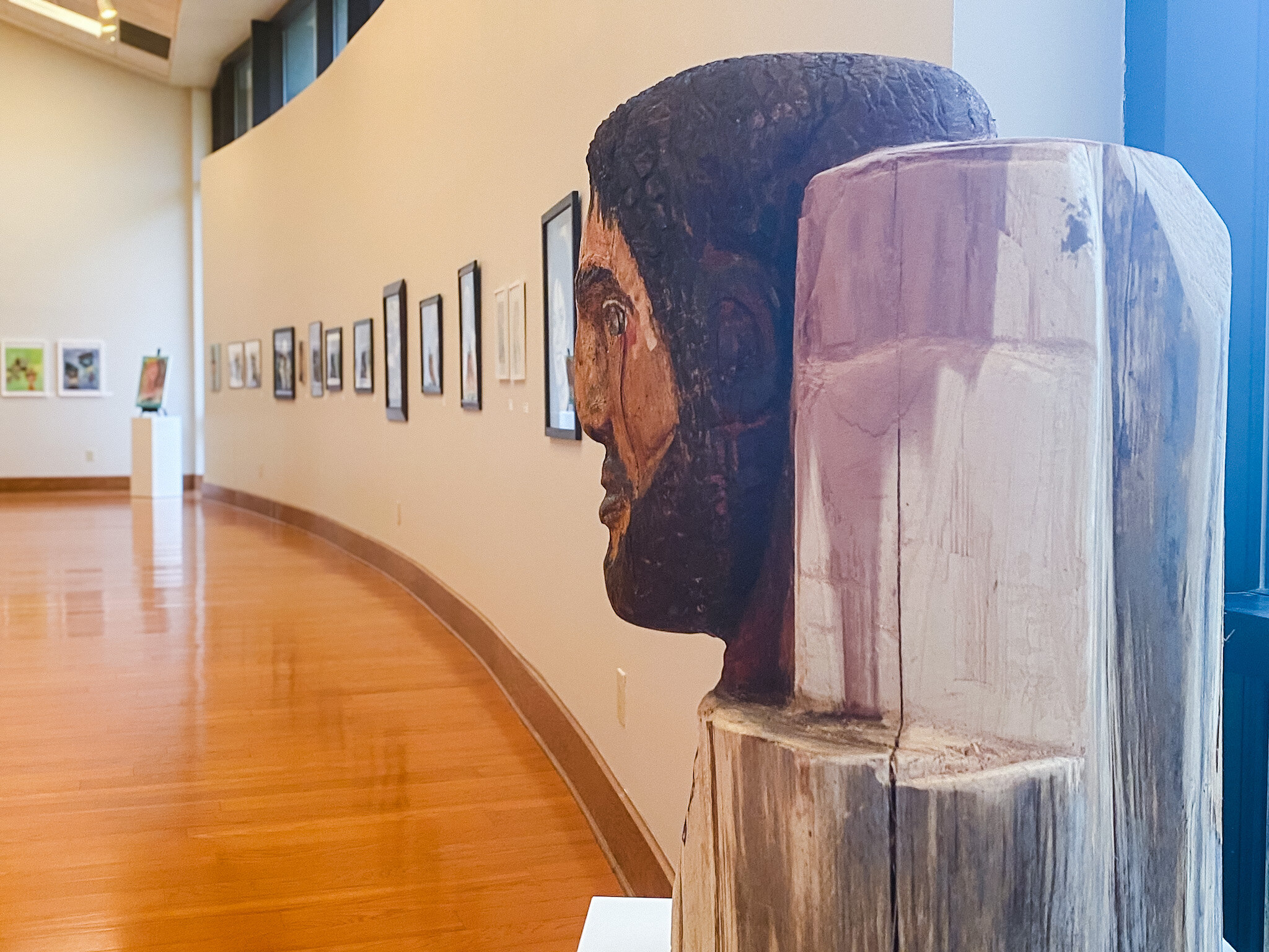Wooden sculpture of a man's head on a pedestal in an art gallery with polished floors and framed art along a curved wall.