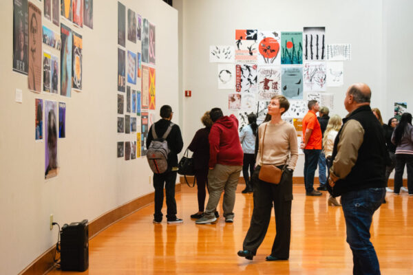 People discuss colorful art on white walls in a brightly lit gallery with a polished wooden floor.