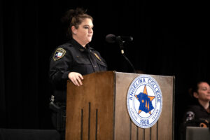 Police officer speaks at podium with Angelina College seal; another uniformed person sits on stage in the background.
