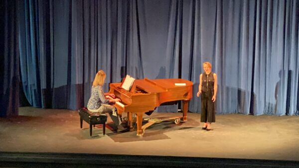 Woman plays grand piano on stage as another woman sings beside her, both performing before closed blue curtains and stage lights.