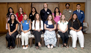 Fifteen young adults in semi-formal attire pose indoors, eight seated and seven standing, all smiling with wood wall behind.
