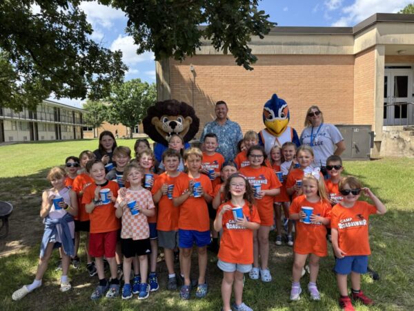 Children in orange Camp Roadrunner shirts pose with two adults, a lion mascot, and a blue bird mascot outside a school building.