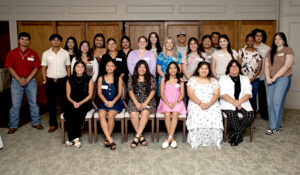 Twenty-three young adults in semi-formal wear pose indoors, seated and standing, smiling in a bright room with wooden paneling.