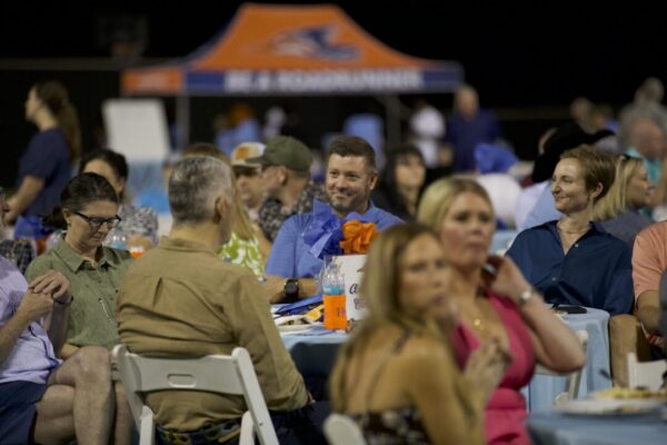 People sit at outdoor round tables, eating and socializing at an event. A tent with blurred lettering is in the background.