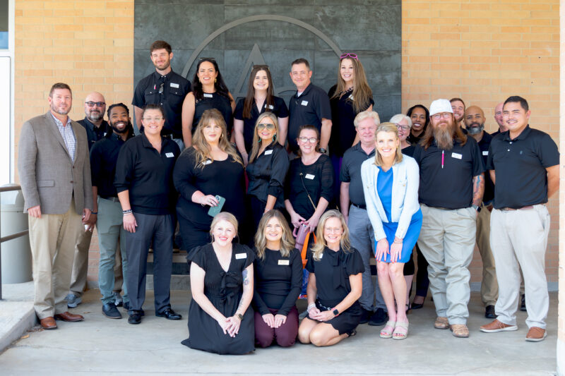 Twenty-two adults in mostly black outfits pose and smile by a brick wall with a large circular logo; four women kneel up front.