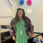 att.VdlOsjaLaLab4QiKKr99BxHaAiGWaC1w9g5av3vHxt0 Smiling woman with curly hair in a green dress and floral kimono stands in a colorful classroom, “You Got This” sign behind her.