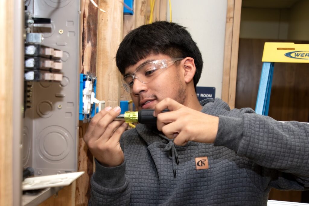 Young man in safety glasses and gray hoodie uses a screwdriver on panel wiring; yellow ladder and wooden wall in background.