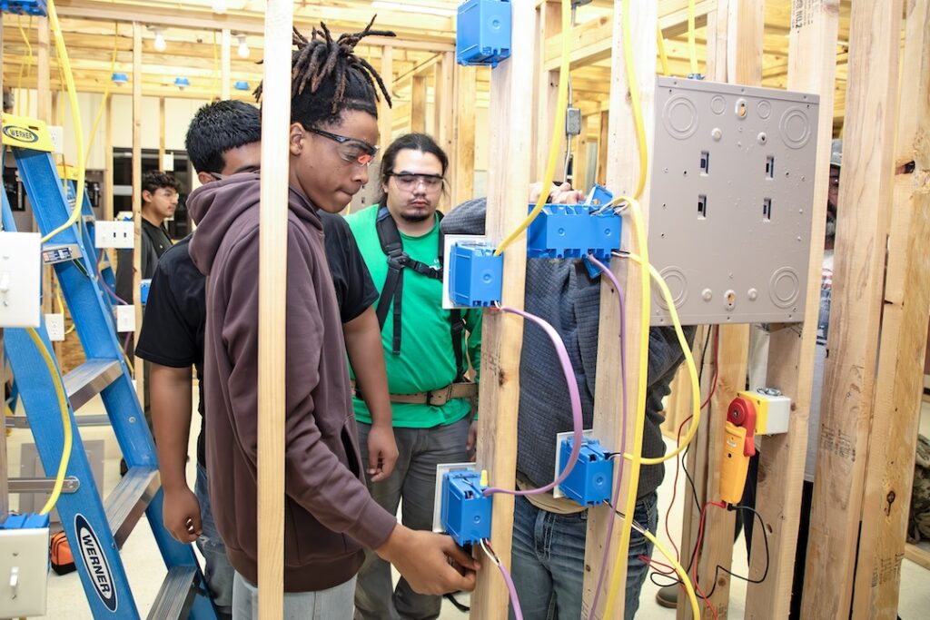 Students in safety glasses wire electrical outlets and switches in a building under construction with exposed framing.