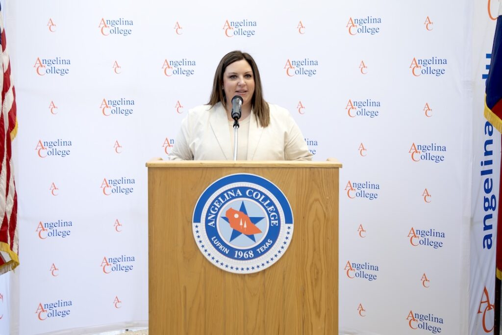 Woman speaks at a podium with the Angelina College logo, backed by a branded banner and partial view of an American flag.