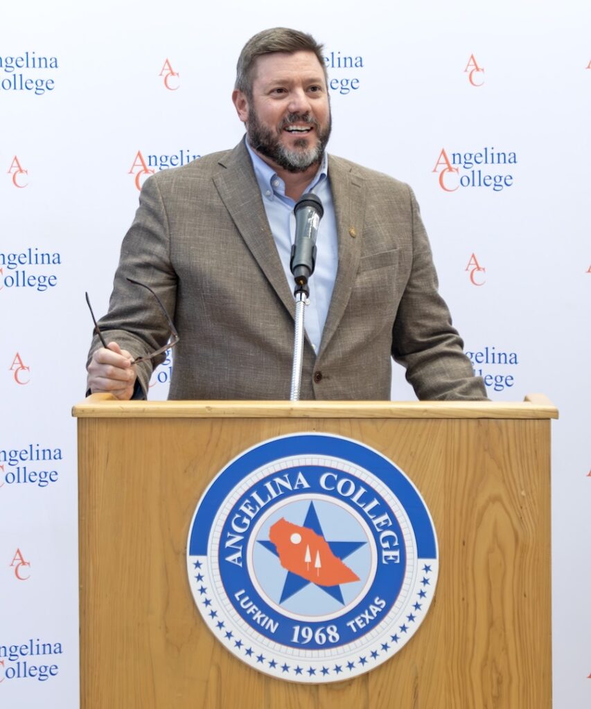 Man in a brown blazer speaks at an Angelina College podium, holding glasses; backdrop has repeated college logos and text.