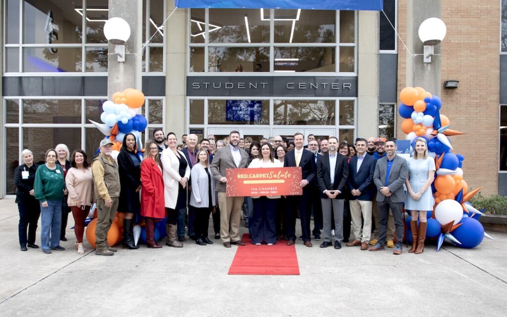 Group poses on red carpet at Student Center entrance, flanked by orange and blue balloons, holding ribbon for ceremony.