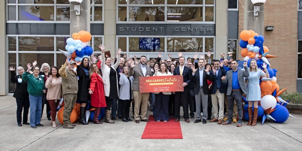 People gather, smiling and cheering, behind a "Red Carpet" ribbon outside the Student Center, with balloons at the entrance.