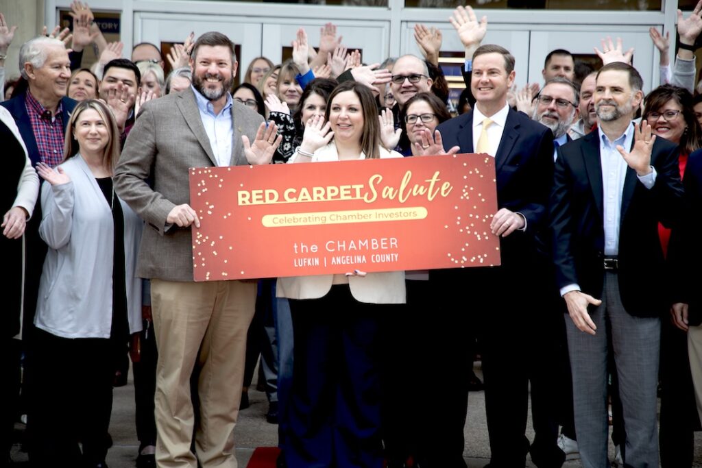 Professionally dressed group smiling and waving behind a sign reading Red Carpet Salute: Celebrating Chamber Investors.