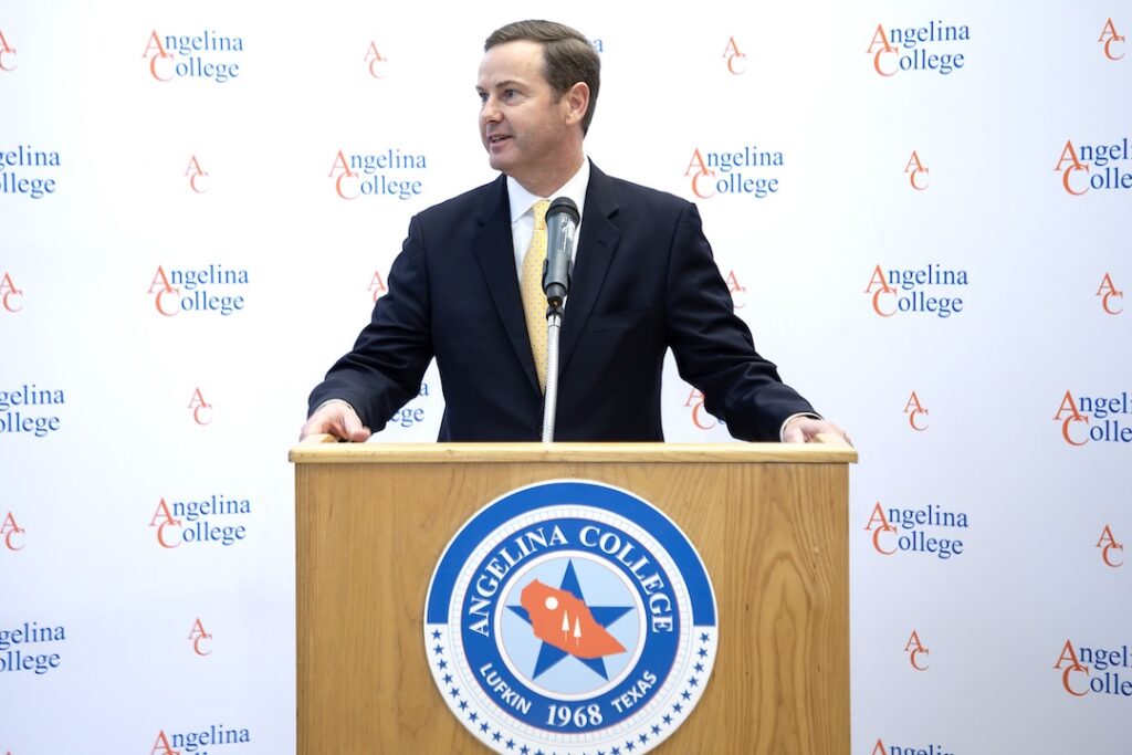 A suited man speaks at a podium with the Angelina College logo, in front of a backdrop with repeated college logos.