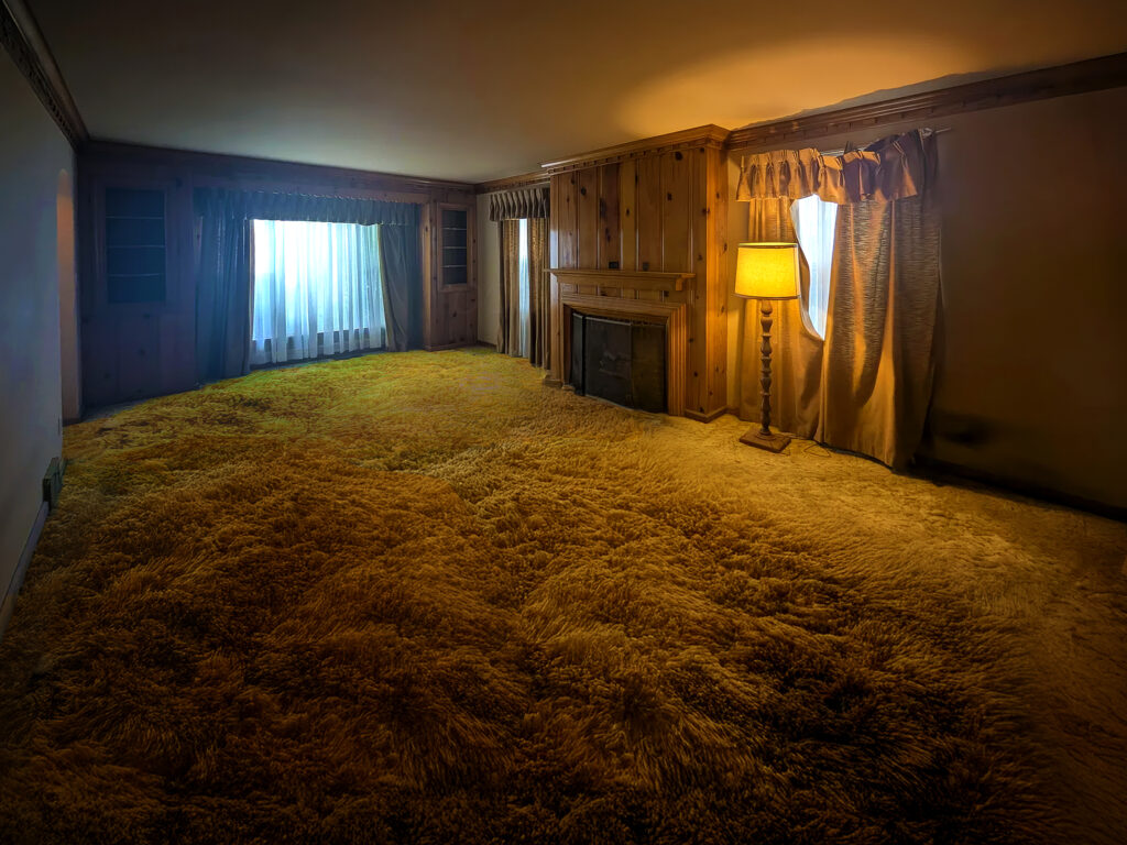 The photo is an example of the work from artist/photographer Chris Ireland. It is a photograph of a living room without furniture with a fireplace and lamp and shag carpet. 