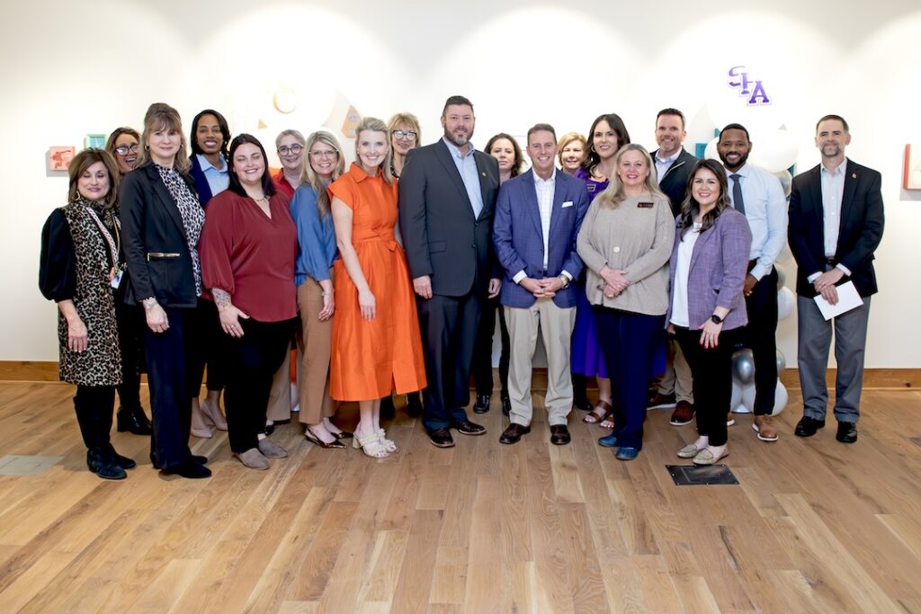 Angelina College and Stephen F. Austin State University staff stand together for a group photo at the Jacks Jumpstart signing event, inside a gallery space with balloon columns and artwork on the walls.