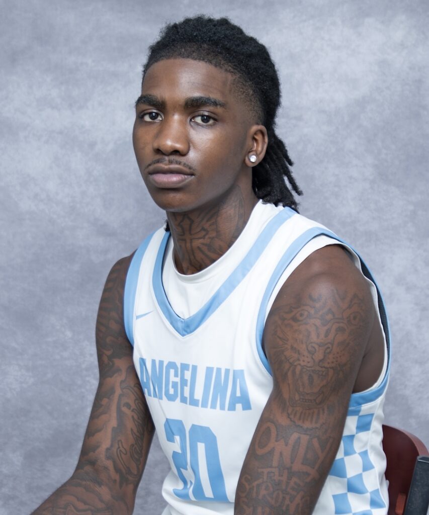 Young male basketball player with tattoos, earrings, and dreadlocks wears ANGELINA 30 jersey, seated against gray background.