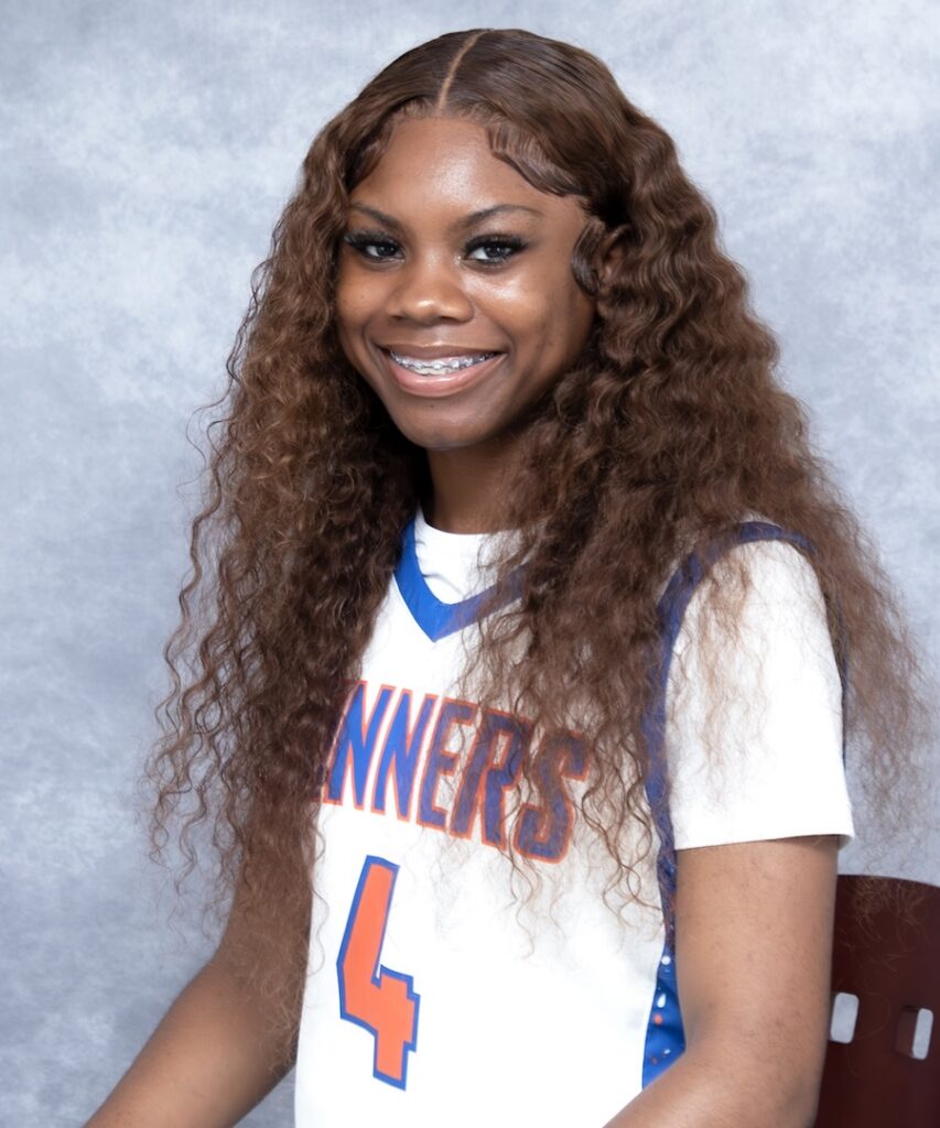 Smiling young woman with long curly brown hair in a white basketball jersey (#4), seated on a maroon chair, gray background.