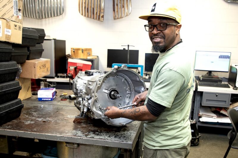 Man in glasses and yellow cap repairs car transmission at metal workbench, surrounded by tools, boxes, and monitors in workshop.